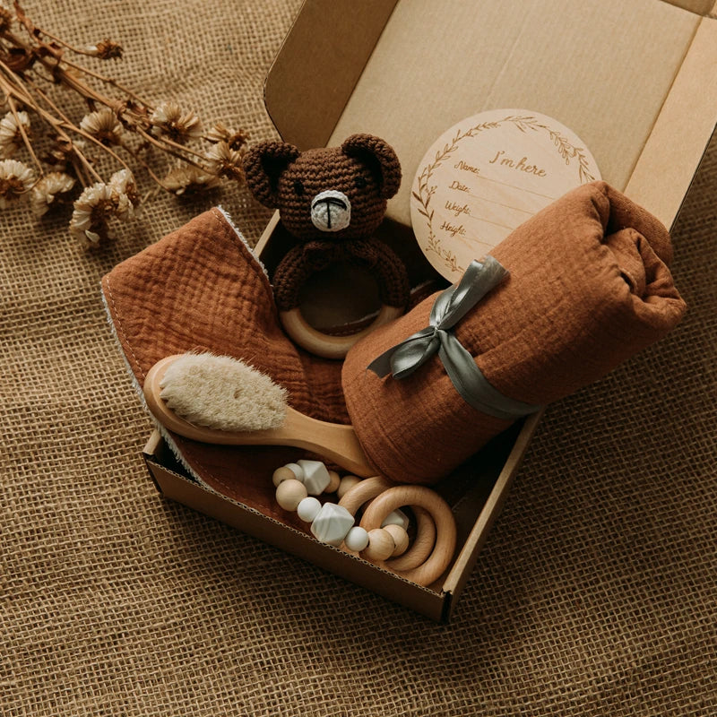A gift box with a knitted teddy bear rattle, brown swaddle, wooden baby brush, wooden teething rings, and a round name plaque. Dried flowers and burlap surface in the background.