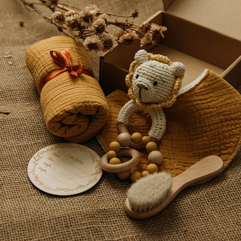 Baby gift set on burlap, featuring a rolled mustard blanket with orange ribbon, a crochet lion rattle, a wooden teething ring with beads, a brush, and a name card.