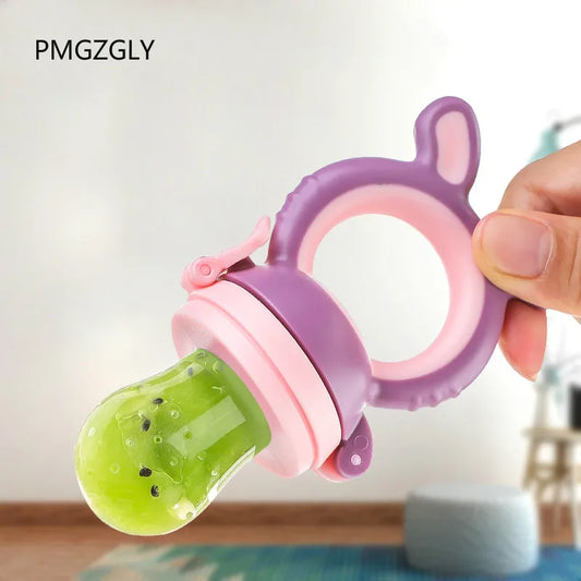 Close-up of a hand holding a pink and purple silicone baby food feeder with a green food-filled sac, designed for safe feeding. The background is a softly blurred room.