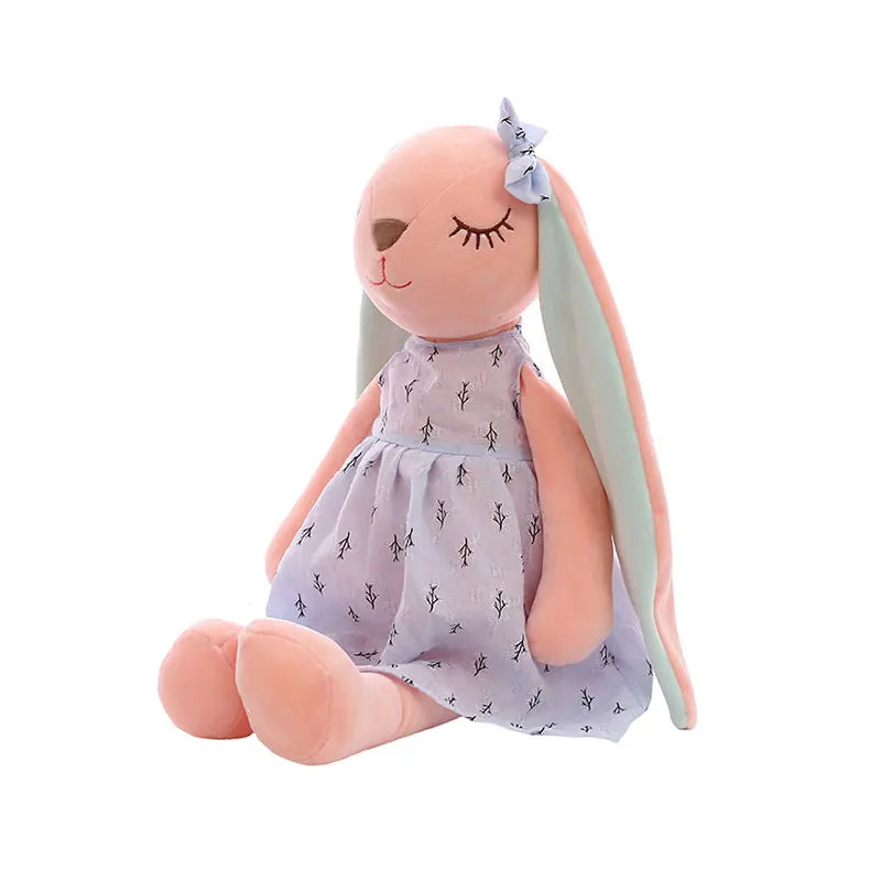 Soft plush pink bunny with long ears and closed eyes, wearing a light purple dress with tiny leaf patterns and a matching bow on one ear, sitting upright.