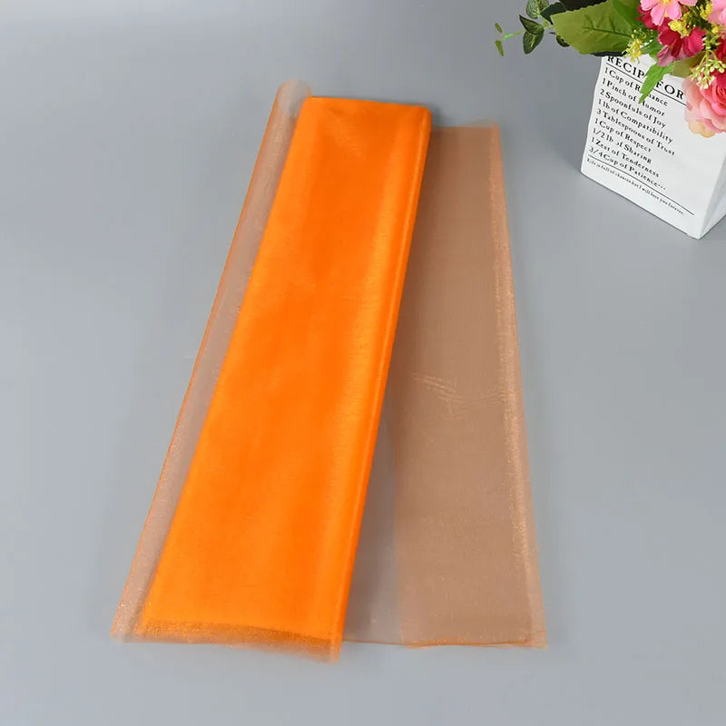 Two sheets of mesh fabric, one bright orange and the other peach, lie on a gray surface. A bouquet of pink flowers and a white box with text are partially visible.