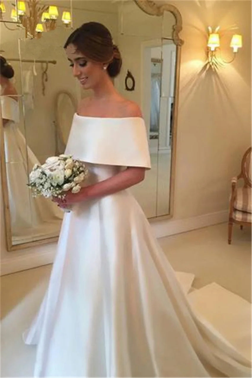 Bride in an elegant off-shoulder wedding gown holds a bouquet of white flowers. She stands in a beautifully lit room with a mirror and soft lighting, exuding elegance and joy.