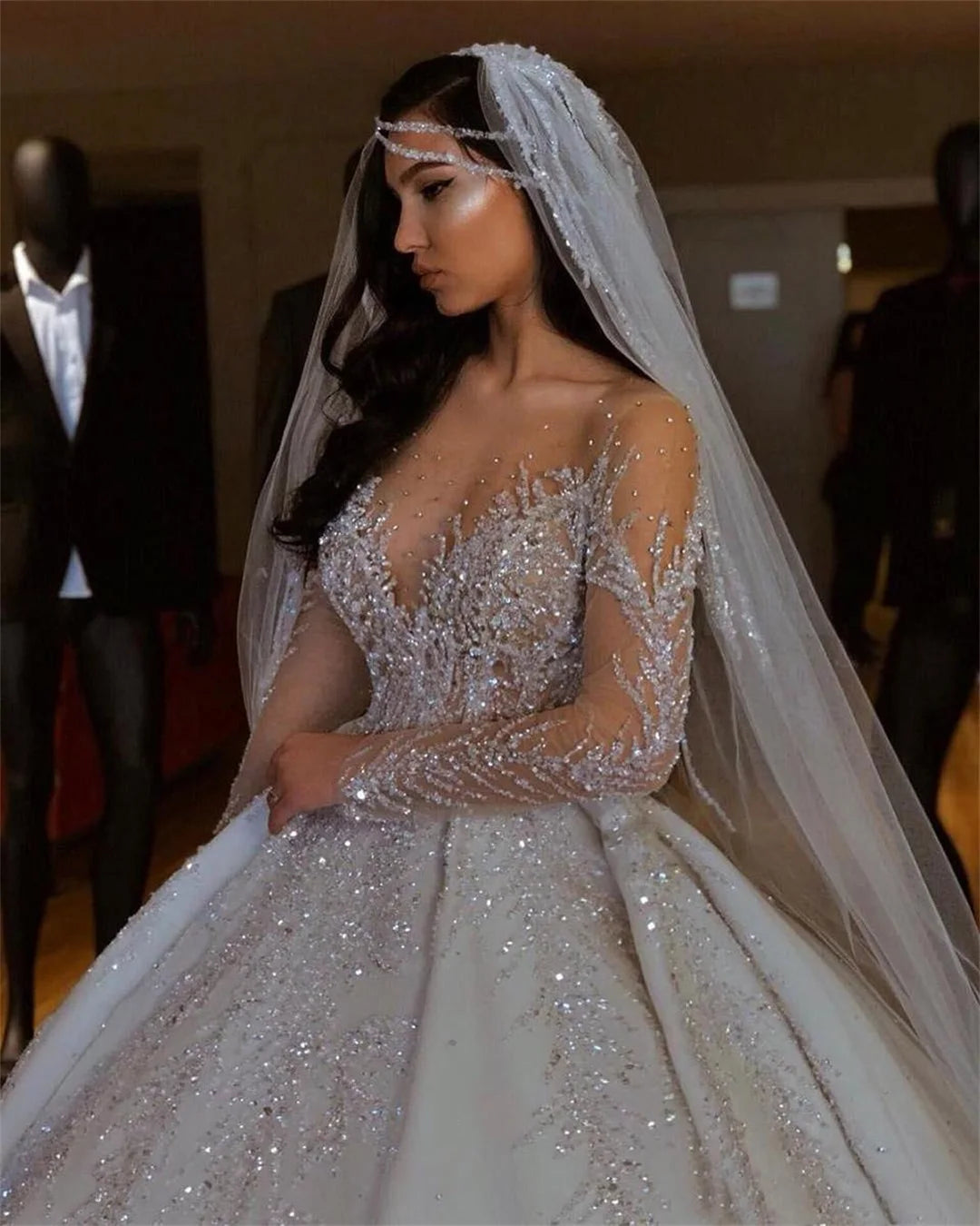 A bride in a sparkling, intricately beaded wedding gown with sheer long sleeves and a bejeweled headband. Her veil cascades, complementing the dress's embellishments.