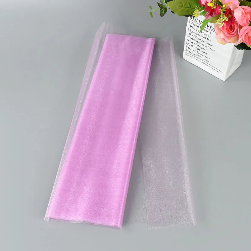 A folded piece of pink organza fabric lies partially wrapped in transparent fabric on a gray surface. Nearby, a bouquet of pink flowers is in a white paper vase.