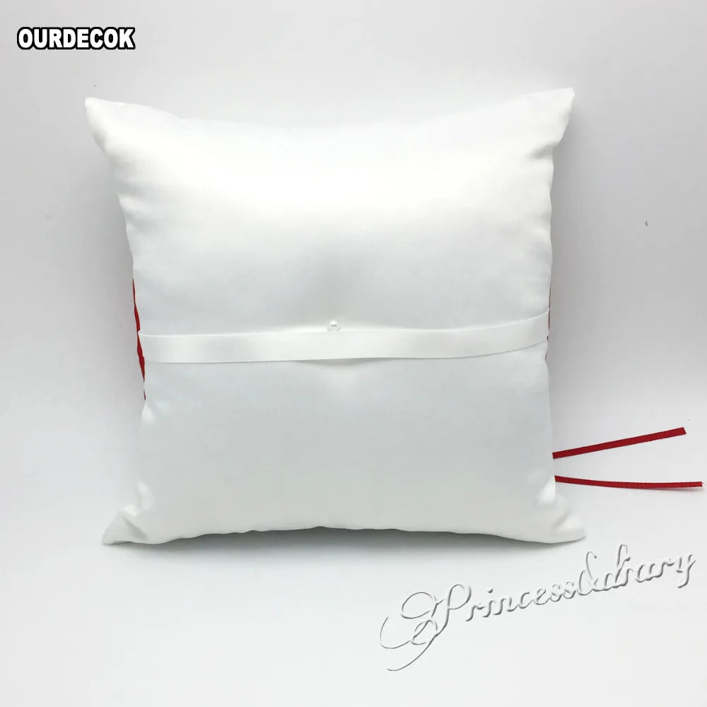 A white decorative cushion featuring a satin ribbon with a small button in the center. Red accents are visible on the side. The text "OURDECOK" and "PrincessDiary" is displayed.