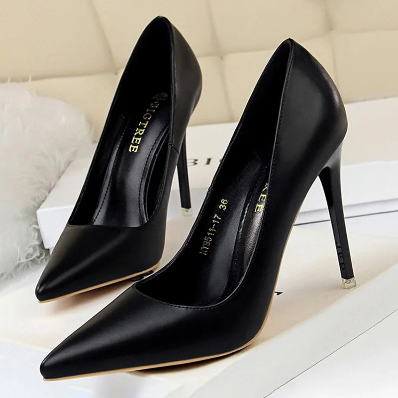 Sleek black high-heeled pumps with a pointed toe and thin stiletto heel, resting on a cream-colored surface with soft, fluffy fabric nearby.
