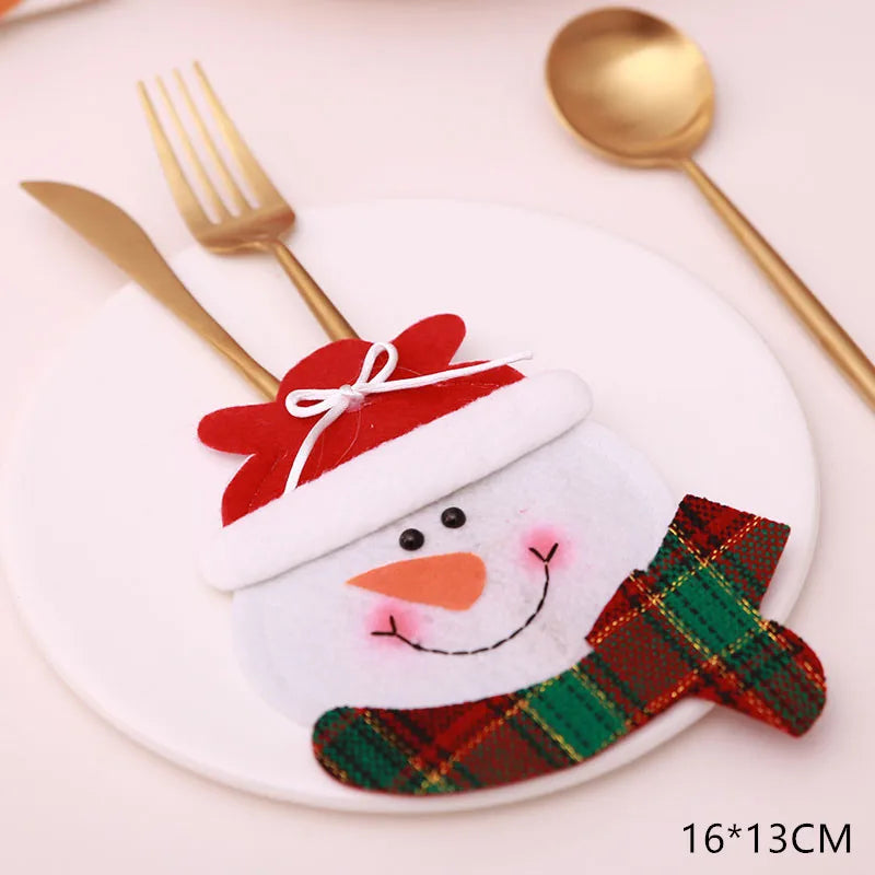 A festive snowman cutlery holder with a red hat and green plaid scarf decorates a round white plate. Gold fork and spoon are placed inside. Image shows dimensions 16x13 cm.