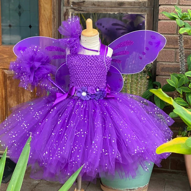 Child-sized mannequin dressed in a vibrant purple fairy costume with wings, adorned with glittery tulle, flowers, and ribbons, set against a wooden and brick background with plants.