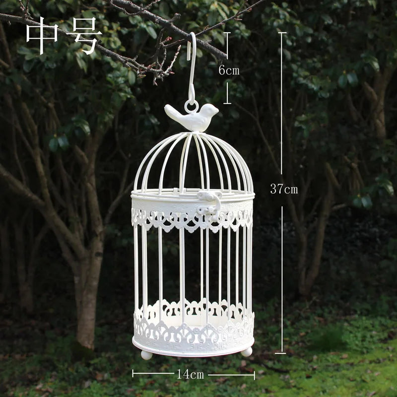 White decorative birdcage with intricate designs, 37cm tall and 14cm wide, hangs from a tree branch outdoors, featuring a bird-shaped ornament on top.