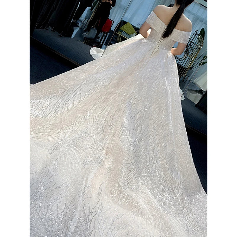 A bride wearing an off-shoulder white gown with intricate sparkling patterns stands with her back to the camera, showcasing the elegant train flowing on the floor.