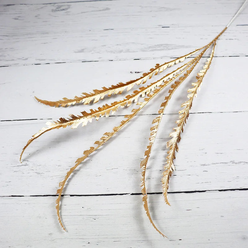 Golden fern-like decorative sprigs with serrated edges lay against a white wooden background, creating a contrast with their shimmering texture.