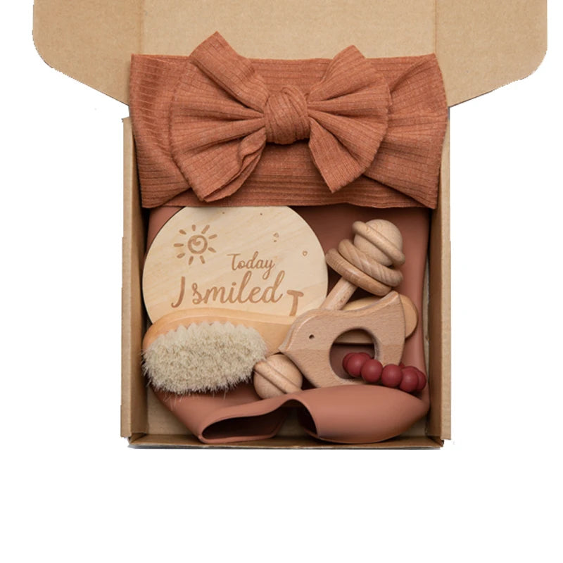 Gift box containing a large bow headband, wooden teether, baby brush with soft bristles, and a round wooden plaque engraved with "Today I smiled," all in a natural brown theme.