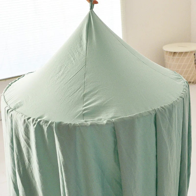 A mint green fabric canopy with a conical top is shown hanging, likely intended as a bed canopy or play tent, in a softly lit room with a round basket in the background.