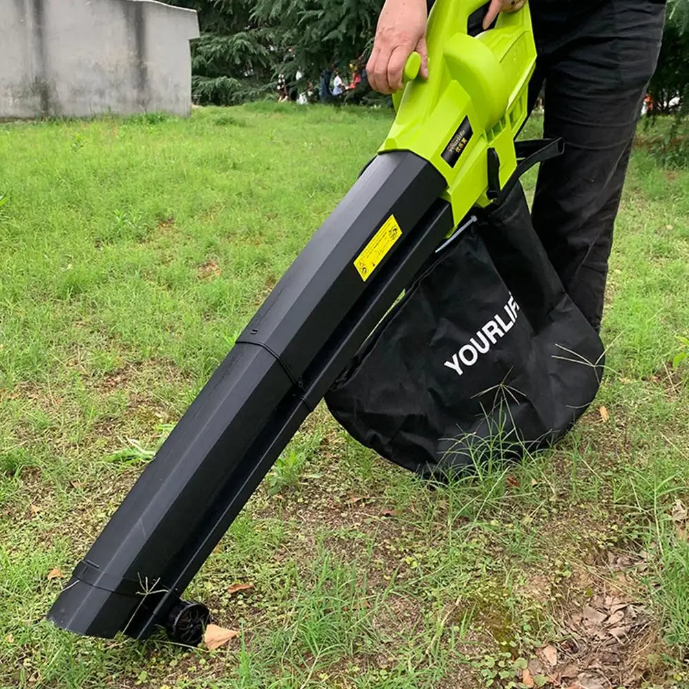 A person uses a green and black leaf blower with a collection bag labeled "YOUR LIFE" on a grassy area, surrounded by trees and a concrete wall.