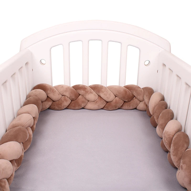 White crib with a braided, plush bumper in shades of brown along the interior edges. The mattress is covered with a light gray sheet.
