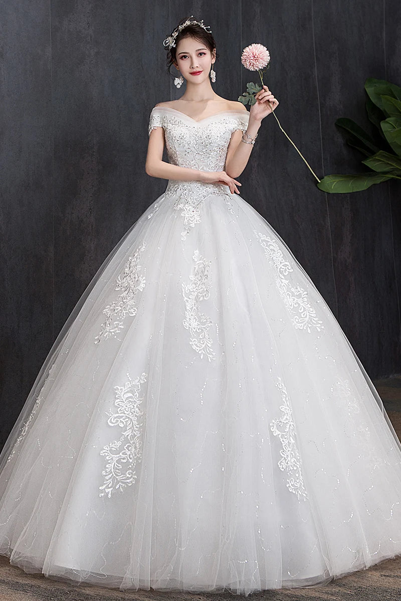 Woman in an elegant off-shoulder white wedding gown with lace details, holding a pink flower. She stands against a dark background with green leaves visible on the side.