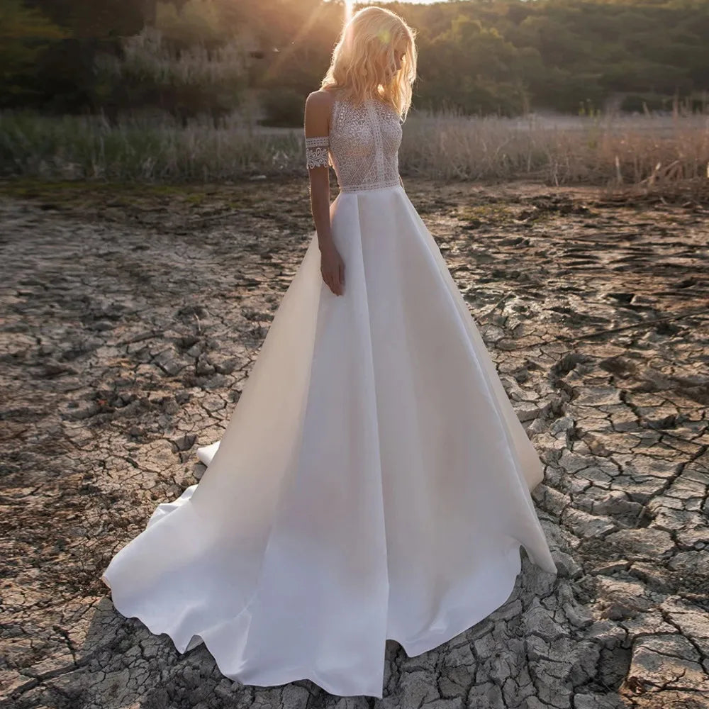 A woman in an elegant white bridal gown stands on dry, cracked earth, with lush greenery and sunlight filtering through trees in the background.