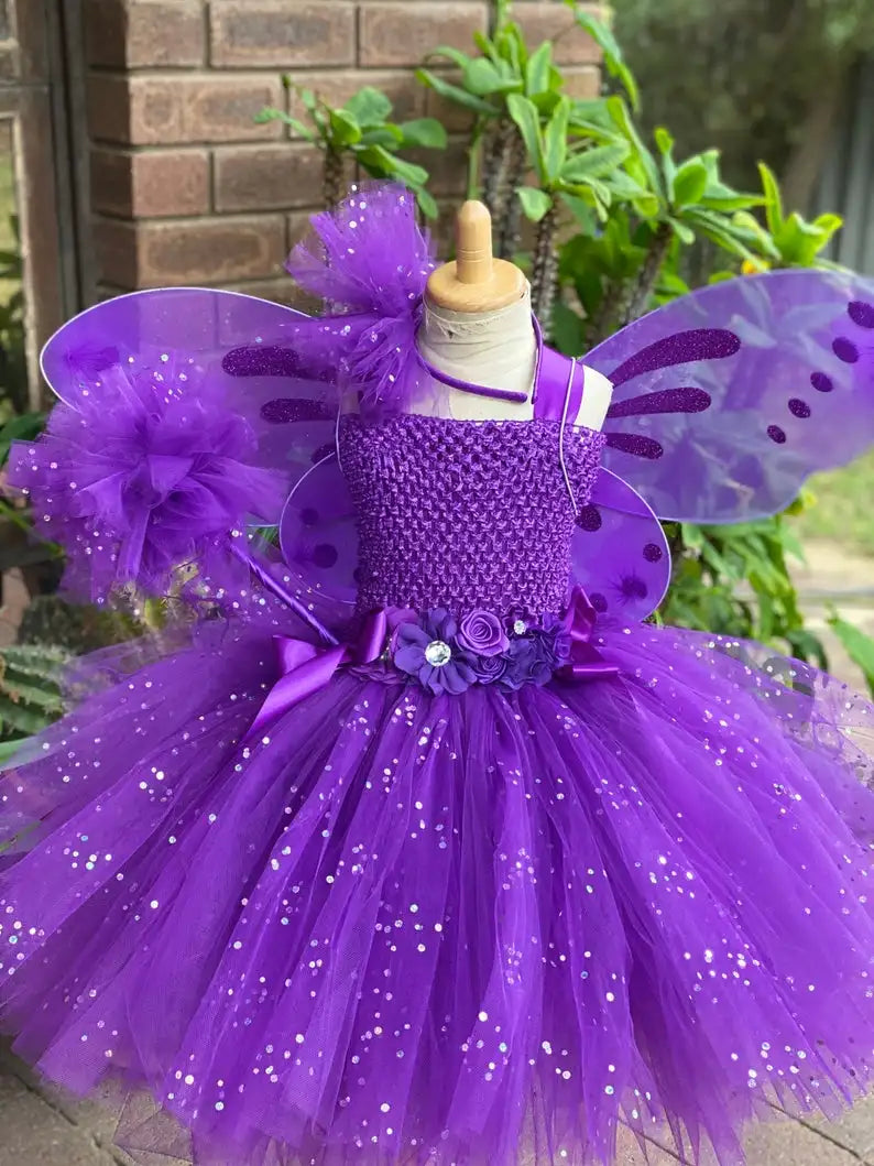 Purple fairy costume on a mannequin with sparkly tulle skirt, floral waistband, and decorative wings. Set outdoors against brick and green foliage backdrop.