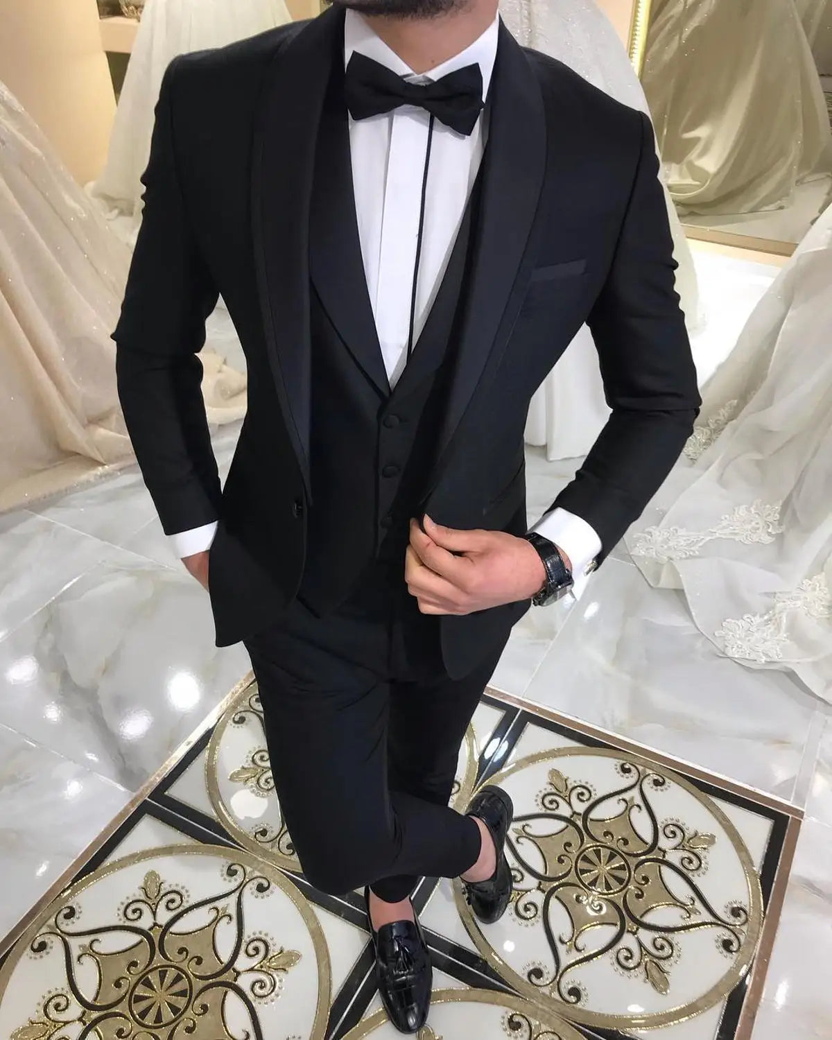 Man in a black tuxedo with a bow tie, hands in pockets, standing on an ornate tile floor. Background features white wedding dresses on display.