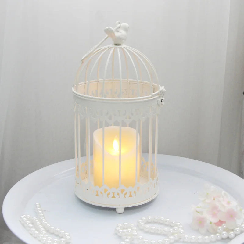 White birdcage with decorative top houses a glowing candle. It's set on a table with pearl strands and pink flowers, creating a delicate, elegant ambiance.