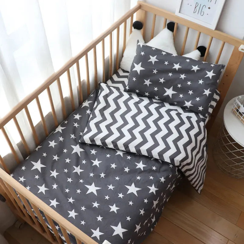Baby crib with wooden rails, star-patterned bedding, and zigzag pillows. A sign on the wall reads "Dream Big." Bright sunlight shines through a sheer curtain.