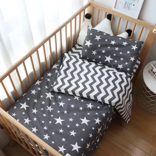 Baby crib with wooden rails, star-patterned bedding, and zigzag pillows. A sign on the wall reads "Dream Big." Bright sunlight shines through a sheer curtain.