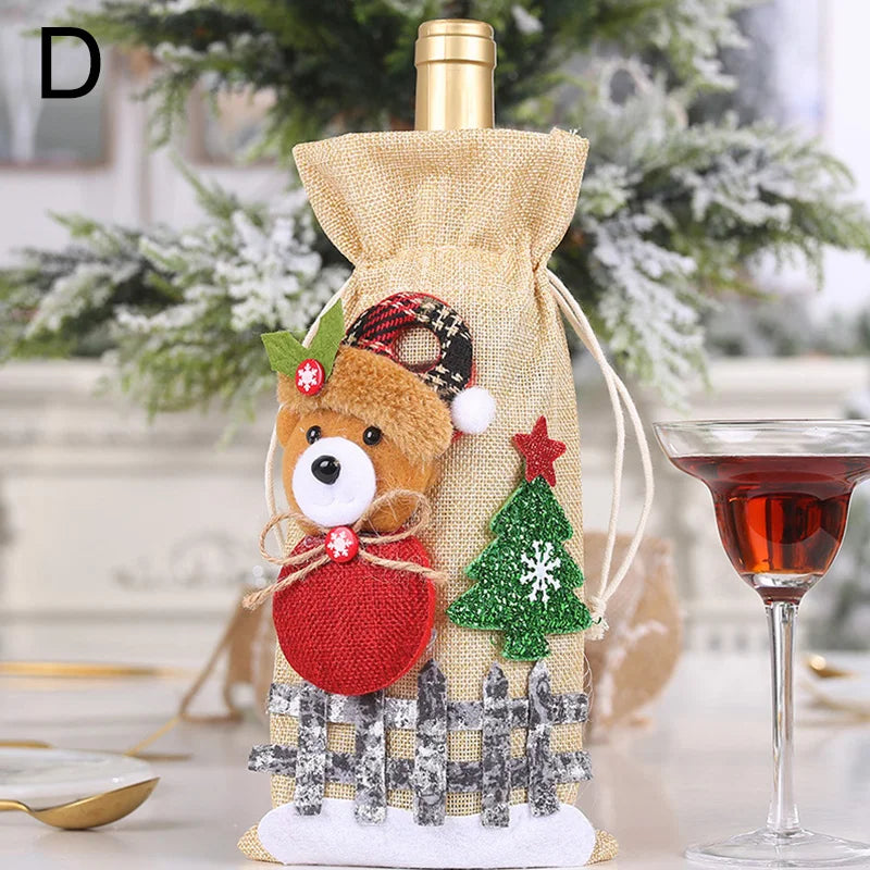 Festive wine bottle in a burlap bag adorned with a bear, plaid hat, red ornament, green tree, and star. Set on a table next to a glass of red wine, with snowy tree backdrop.
