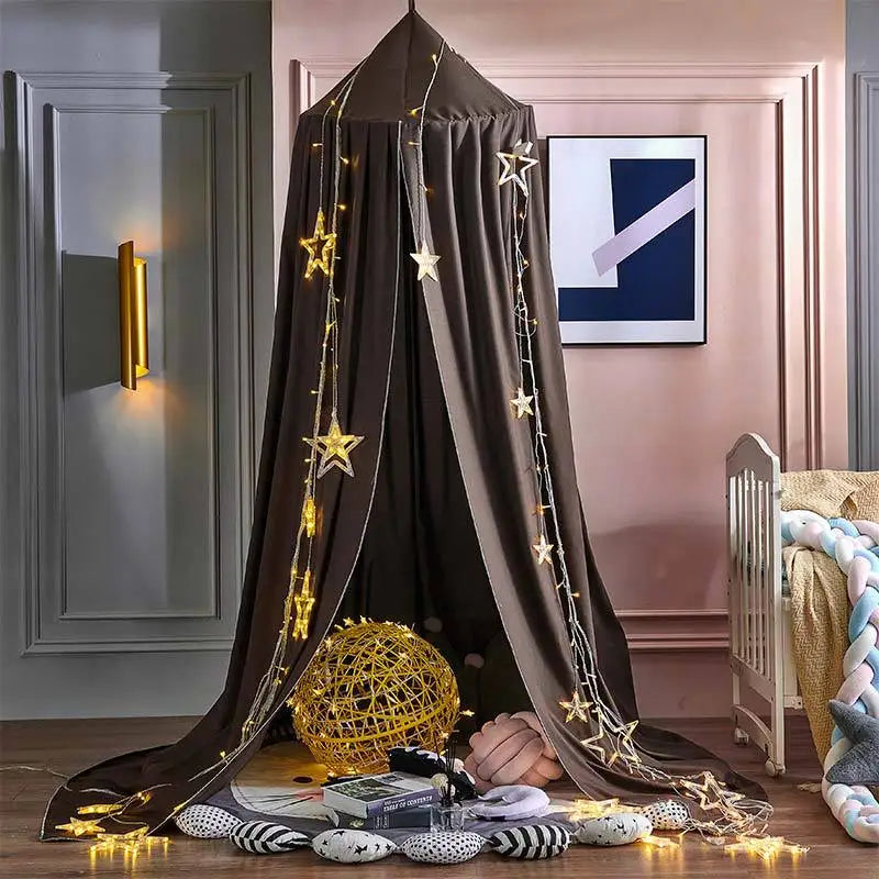 Cozy children's play tent decorated with star-shaped string lights, surrounded by soft cushions, a large woven ball, and a crib in a pastel-colored room with modern wall art.