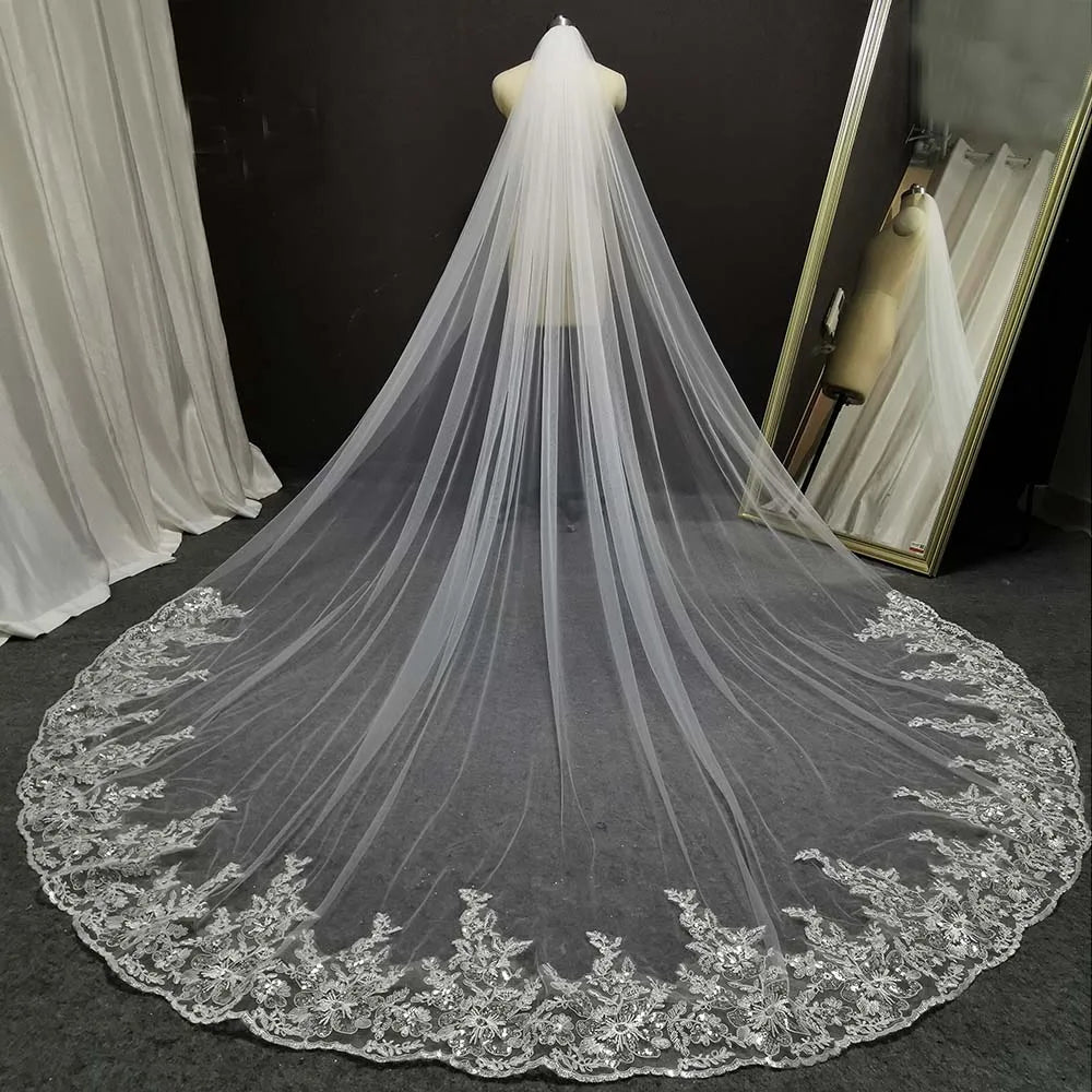 A long, flowing wedding veil with delicate lace embroidery displayed on a mannequin. The veil extends across a gray floor, with a mirror on the right reflecting the scene.