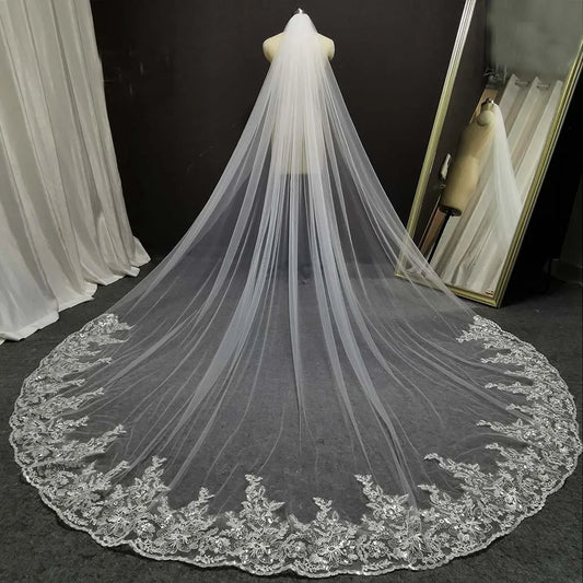 A long, flowing wedding veil with delicate lace embroidery displayed on a mannequin. The veil extends across a gray floor, with a mirror on the right reflecting the scene.
