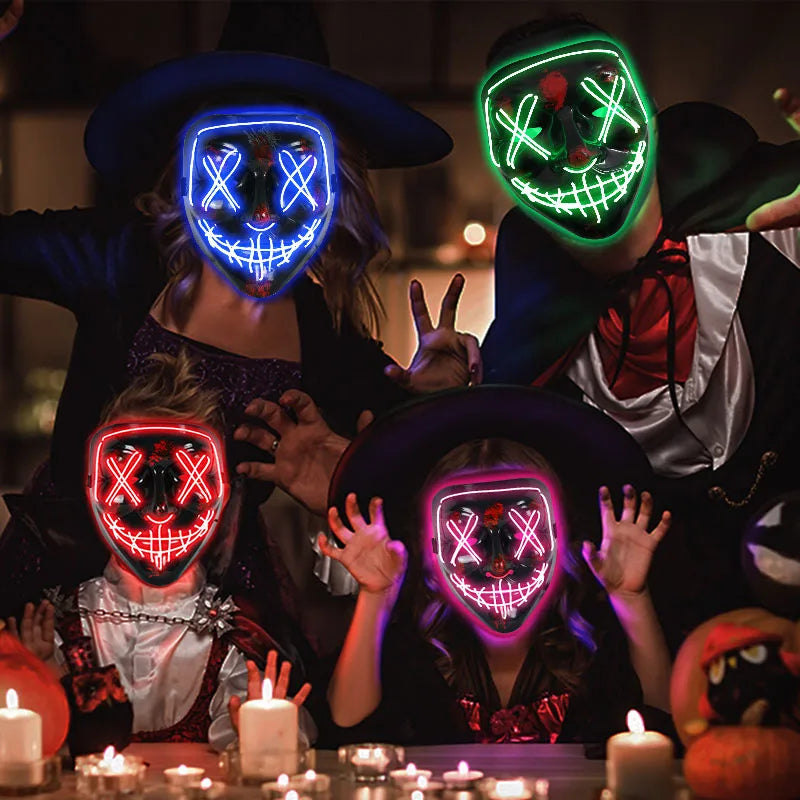 Four people in glowing masks and Halloween costumes gather around a table with lit candles. Masks feature neon X eyes and stitched smiles, creating an eerie, festive atmosphere.