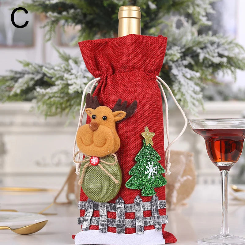 Festive wine bottle in a red cloth bag adorned with a reindeer and Christmas tree decoration, beside a glass of red wine, with a snowy tree in the background.