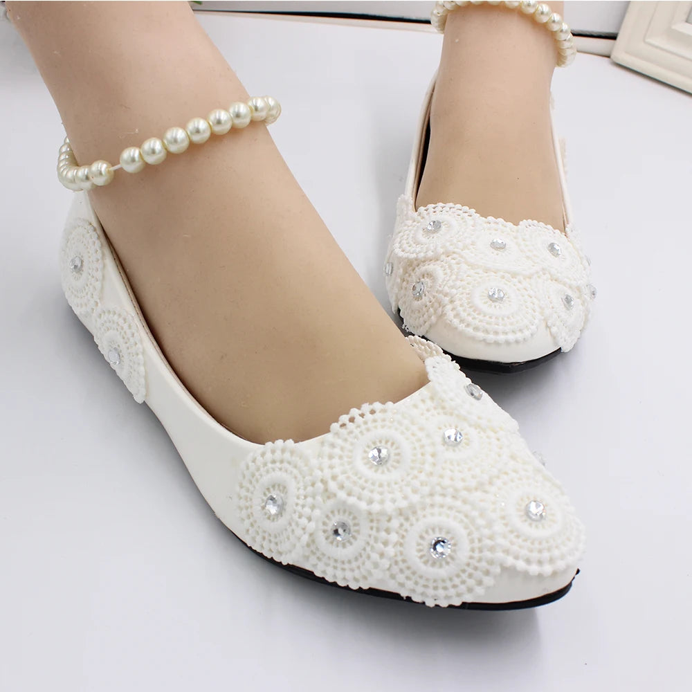 White ballet flats adorned with intricate crochet patterns and rhinestones, worn by feet with pearl anklets, on a smooth white surface.
