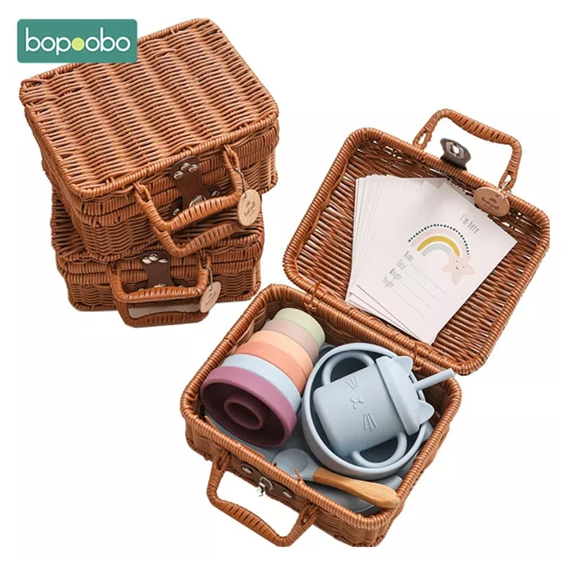 Three woven baskets with handles, one open to reveal pastel-colored silicone toys and a stack of cards featuring a rainbow design. The lid shows the brand logo "bopoobo."