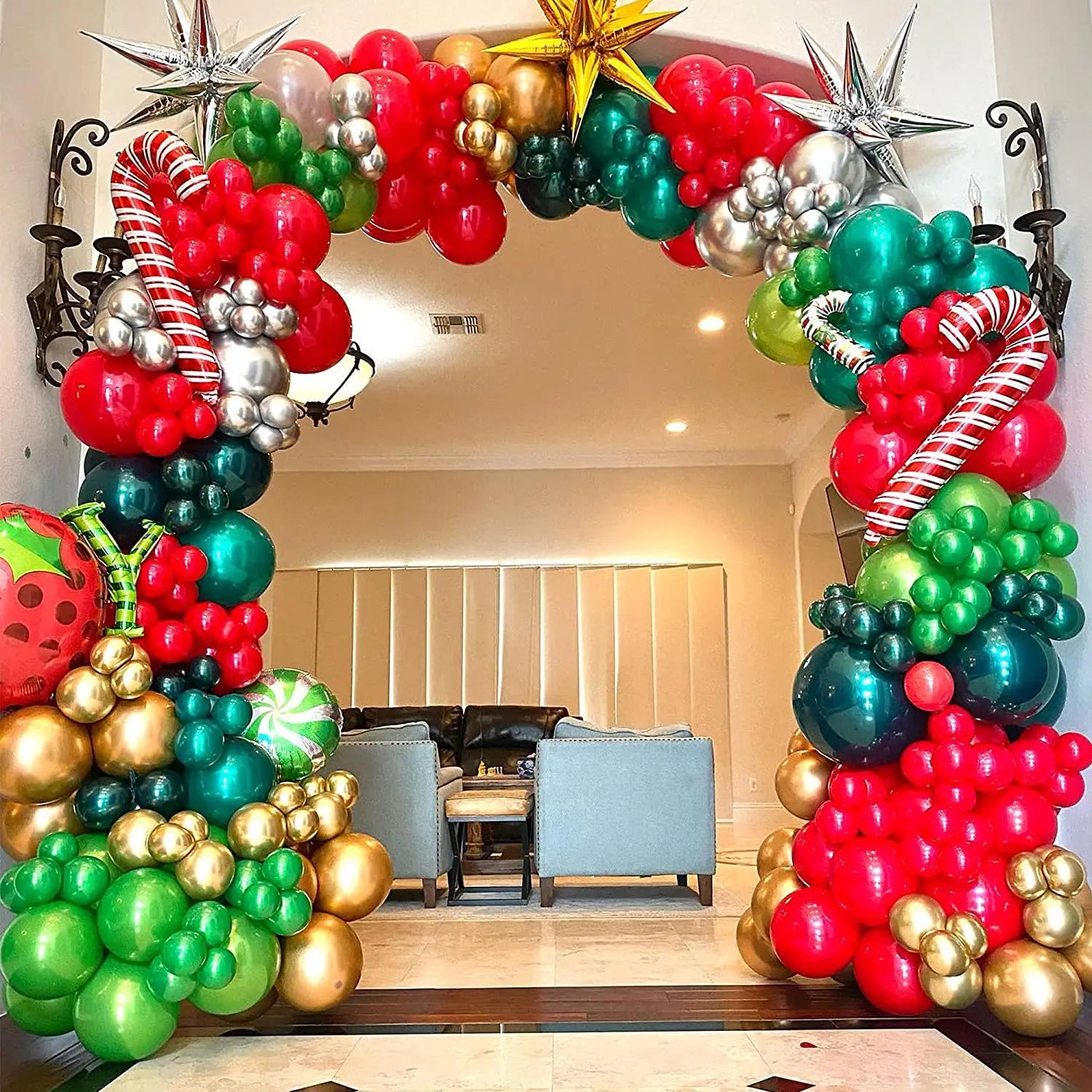 Festive balloon arch with red, green, gold, and silver balloons, candy cane and star details, frames a cozy living room with gray chairs and a sofa, creating a joyful atmosphere.