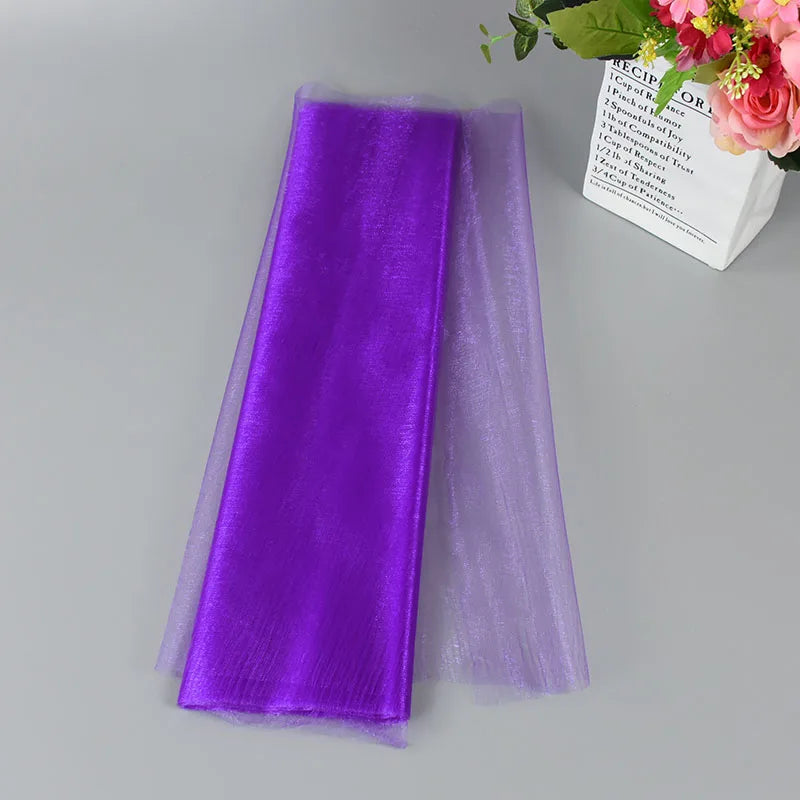 Two folded sheer fabrics, one vibrant purple and the other lavender, lie on a gray surface. A paper bag with colorful flowers is partially visible in the corner.