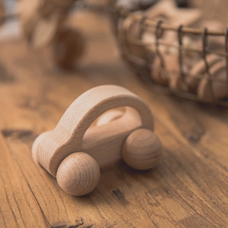 Wooden toy car with smooth curves and round wheels sits on a wooden table. A wire basket filled with similar wooden toys is blurred in the background.