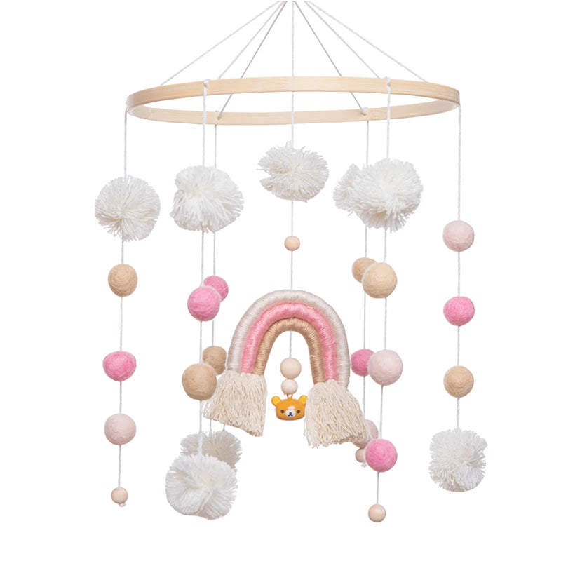 Hanging mobile with white, pink, and beige yarn balls, featuring a central rainbow design with tassels and a bear face, suspended from a wooden hoop.