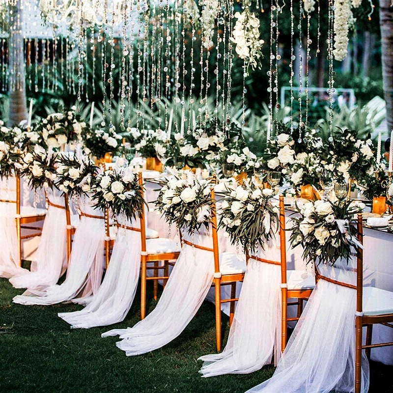 Elegant outdoor wedding setup with long table adorned with white flowers, greenery, and draped fabric on gold chairs. Crystal chandeliers hang above, creating a romantic ambience.