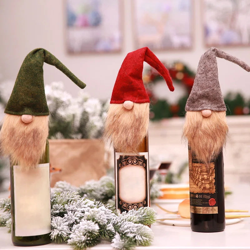 Three wine bottles decorated as gnomes with felt hats and faux beards in red, green, and gray. The background has a festive, blurred holiday decor with frosted foliage.