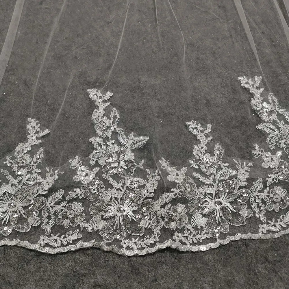 Delicate floral lace featuring intricate flower patterns adorned with sequins and embroidery on a sheer fabric background. The lace lies on a textured gray surface.
