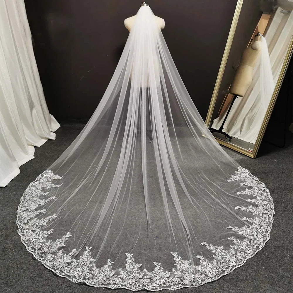 Long white bridal veil with intricate lace edging displayed on a mannequin. The veil extends across a carpeted floor, with a large mirror reflecting its full length.