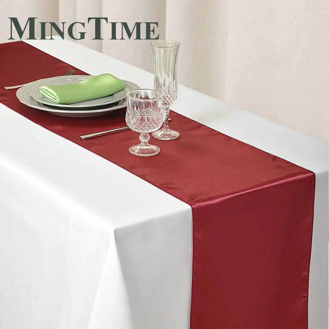 A white tablecloth with a red runner and a place setting, including stacked plates, a green napkin, a fork, and two crystal glasses, one tall and one short.