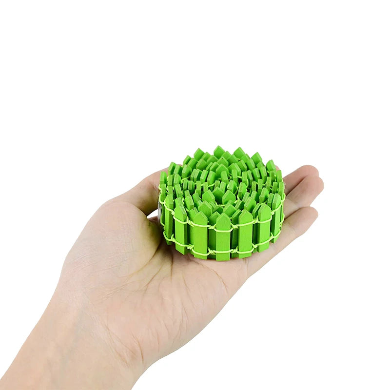 A hand holding a coiled strip of small, bright green plastic fence pieces, resembling a miniature garden fence. The fence is neatly rolled and compact.