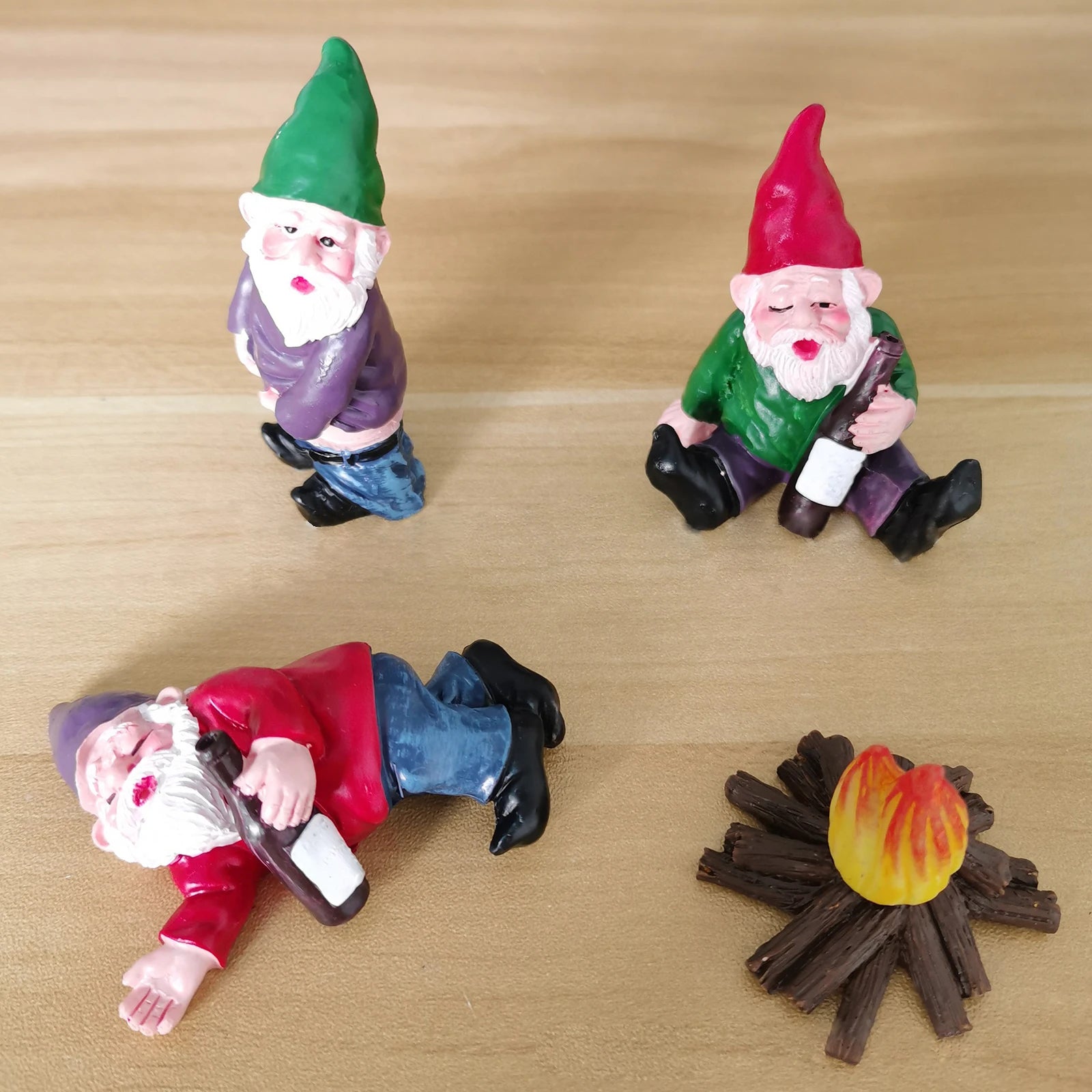 Three decorative gnomes with bottles; one lying down, one sitting, and one standing, near a small campfire on a wooden surface.