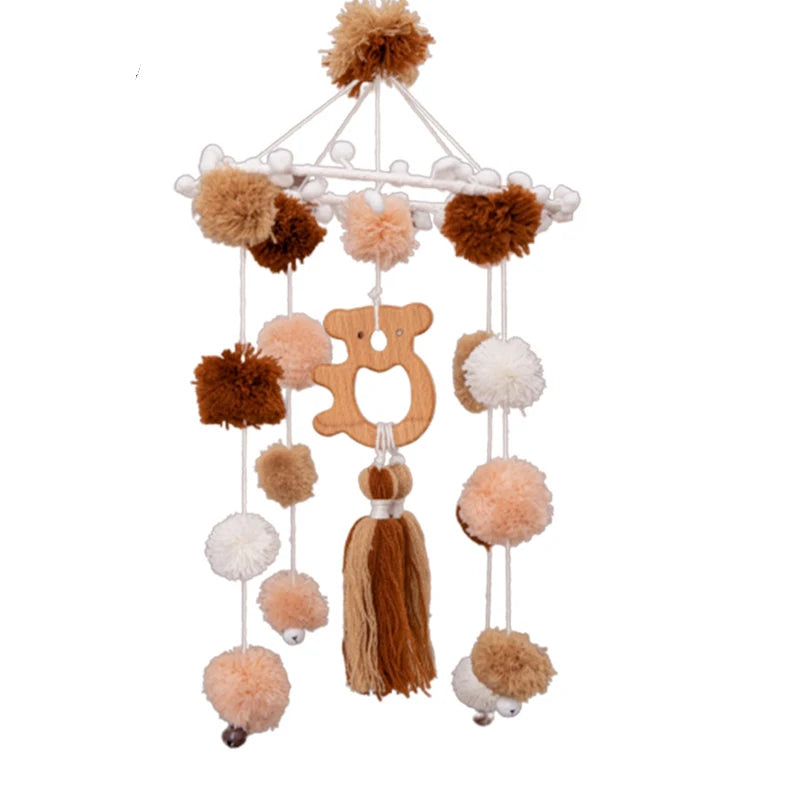 Hanging mobile with brown, white, and beige pom-poms, a wooden bear shape, and tassels. Perfect for a nursery, offering a cozy and playful touch.