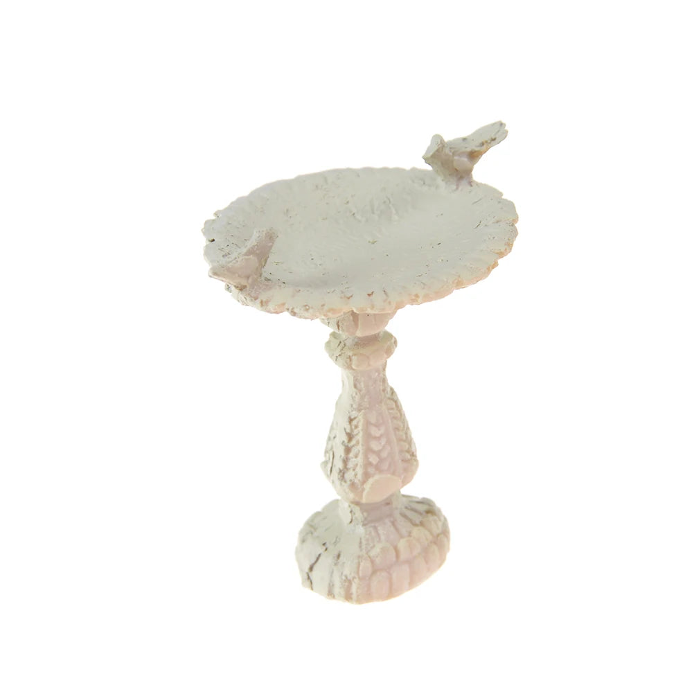 A small, ornate white birdbath with a round basin, featuring intricate details and two tiny birds perched on the edge, set against a plain background.