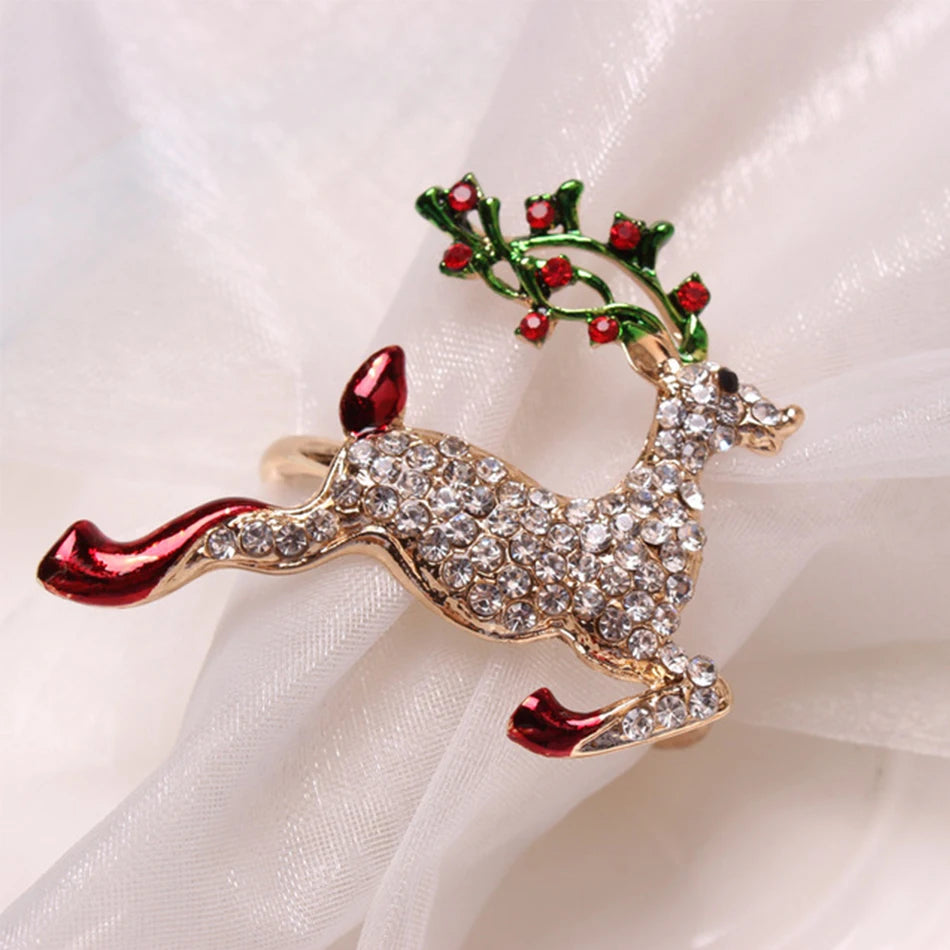 Sparkling reindeer brooch adorned with clear gemstones, green and red antlers, and red hooves, lying on a soft, white fabric background.