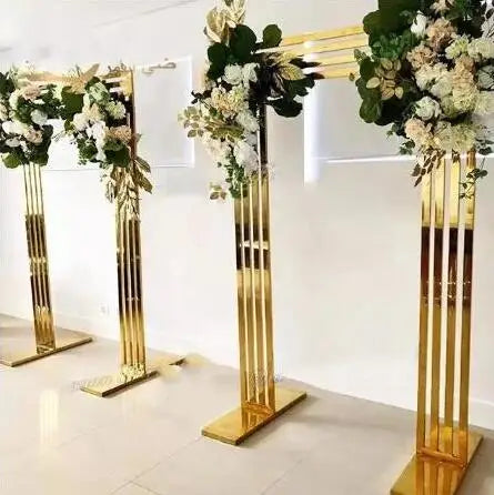 Three gold metal wedding arches adorned with lush white and green floral arrangements, placed in a bright, modern room with a white floor and walls.