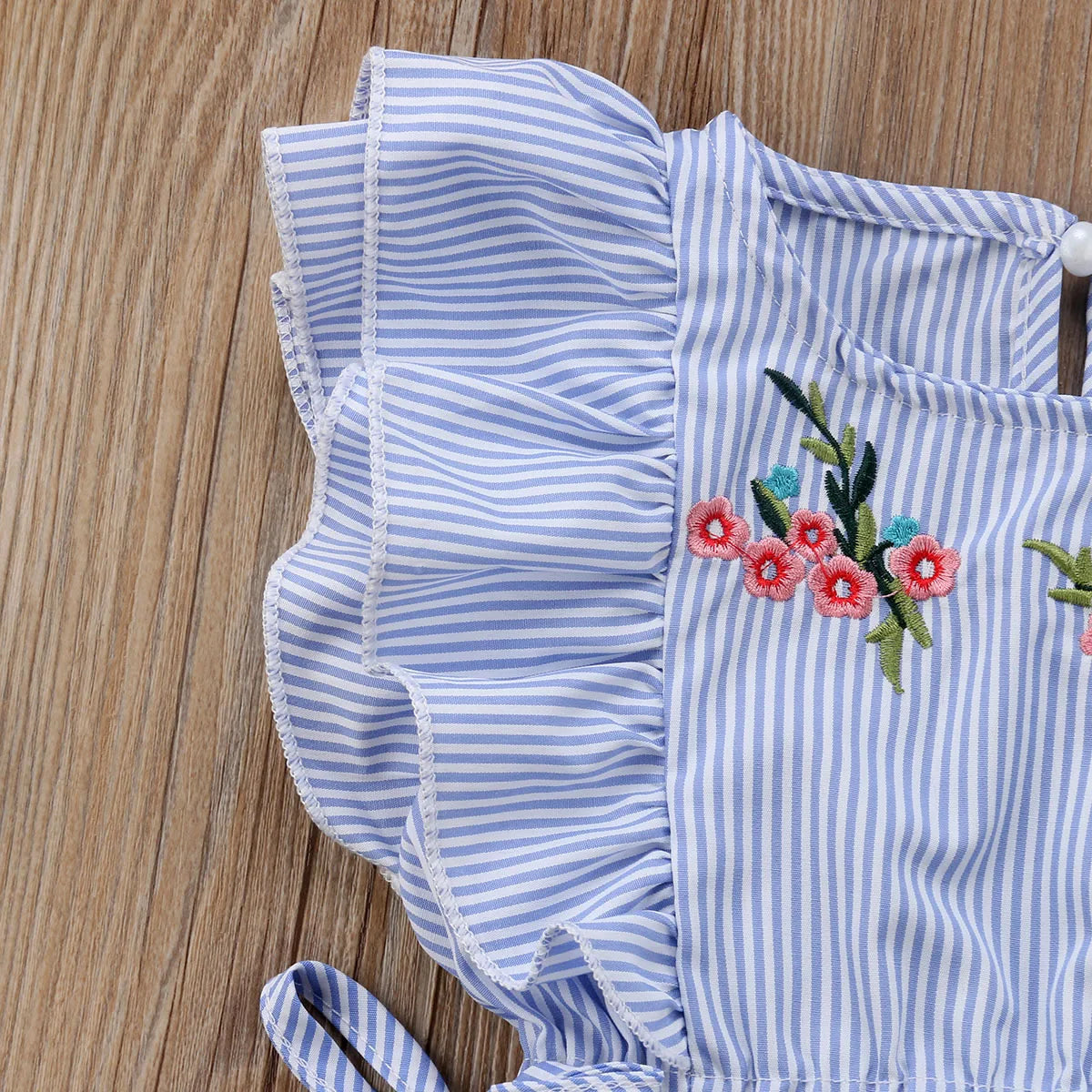 A blue and white striped fabric with ruffled edges, featuring embroidered red and pink flowers with green leaves, lies on a wooden surface.